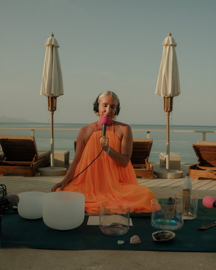 A practitioner in an orange dress leading a sound bath with crystal bowls on a deck above the sea