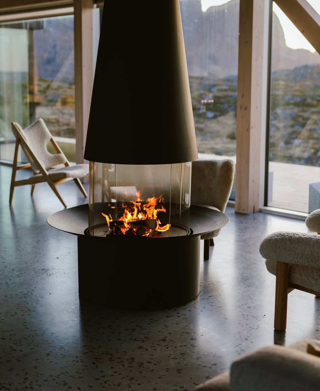 A suspended conical fireplace burning in a lounge with sheepskin chairs and glass walls