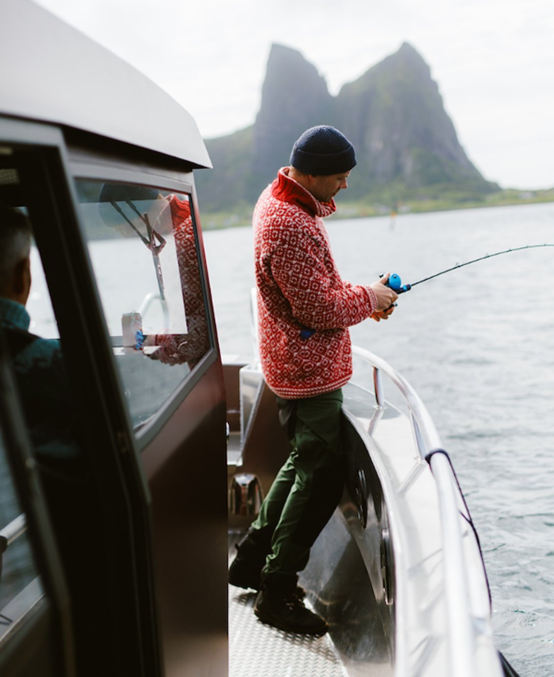 A man in a Norwegian knit sweater fishing from the deck of a boat, a steep peak behind